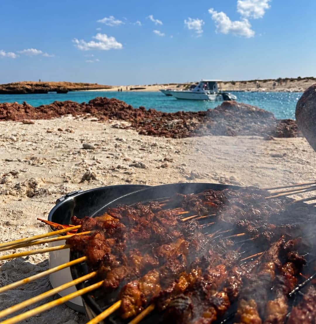Skewers grilling on a Daymaniyat island beach, with the boat anchored offshore, on a full-day snorkelling tour