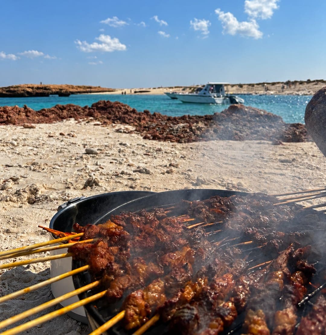 Skewers grilling on a Daymaniyat island beach, with the boat anchored offshore, on a full-day snorkelling tour